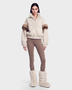 A person wearing the Rudsak ALEIA - 8526693 Vanilla cream faux-fur sweater jacket with fur details, taupe leggings, and cream furry boots stands against a plain white background.