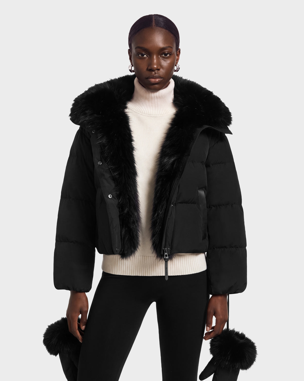A model wears a black Rudsak ALONDRA - 8126992 Black cropped puffer jacket with a large fur collar over a cream turtleneck sweater.