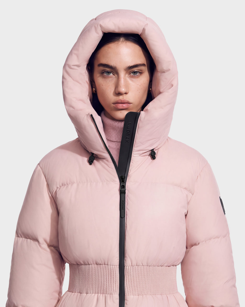A person wearing a pale pink Rudsak DIAMOND - 8126772 Vintage Rose quilted puffer hooded jacket with a cinched waist.