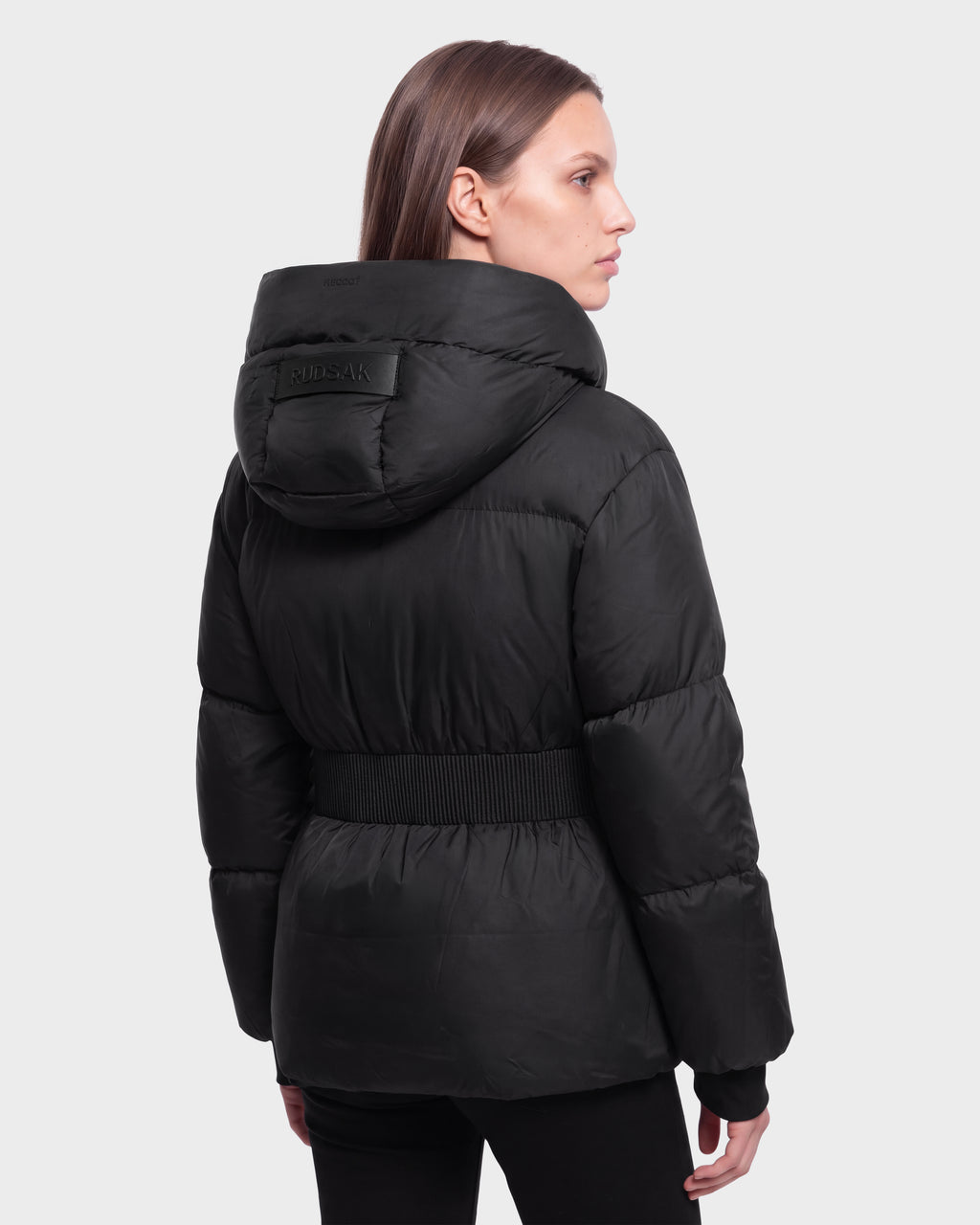 A woman with straight brown hair models the DIAMOND - 8126772 Black by Rudsak, a belted black hooded puffer jacket, standing against a light gray background and facing slightly away from the camera.