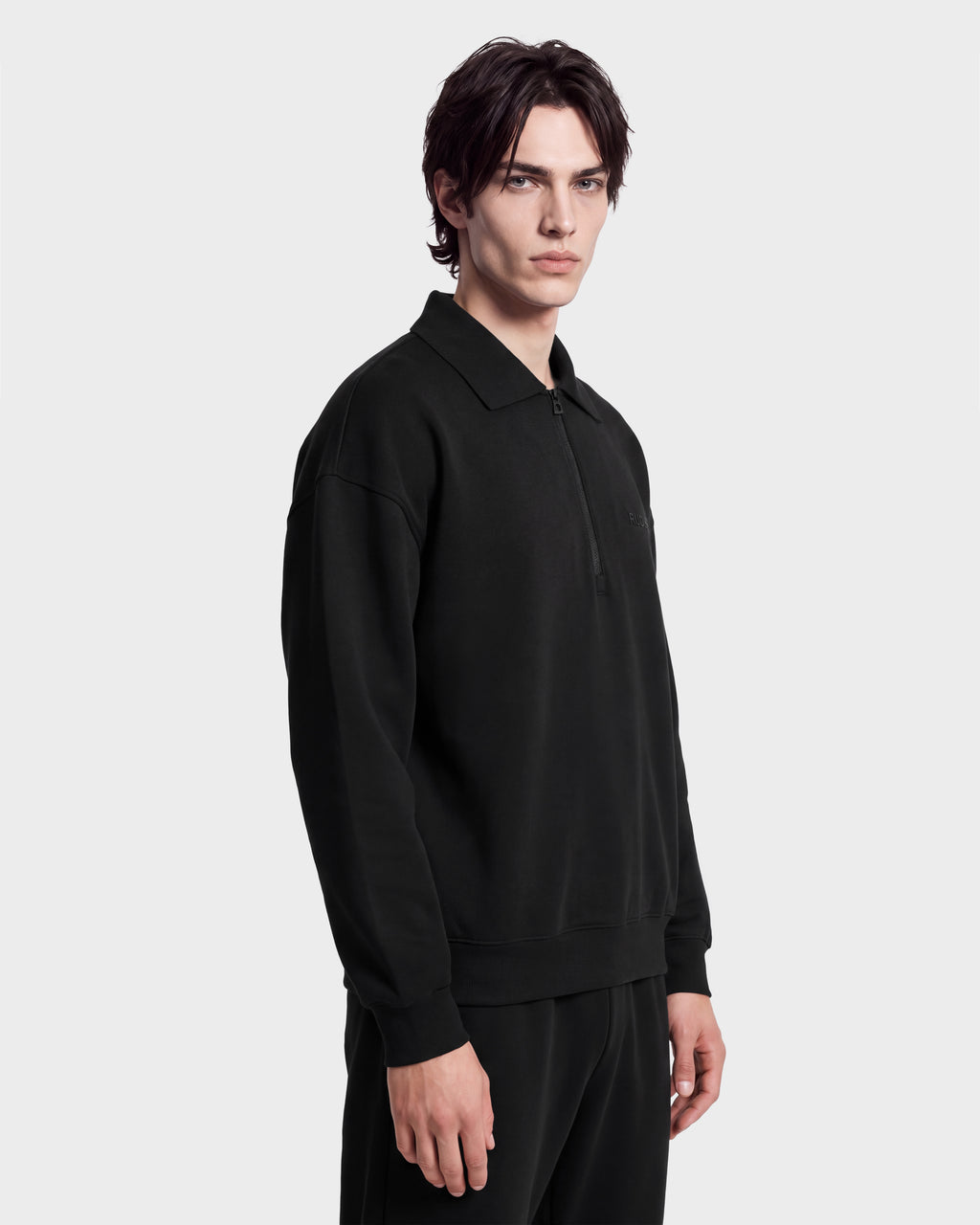 A person with medium-length dark hair models the IDRIS - 6526500 Black by Rudsak, a black collared long-sleeve shirt and matching pants, standing against a plain white background and facing slightly to the side.