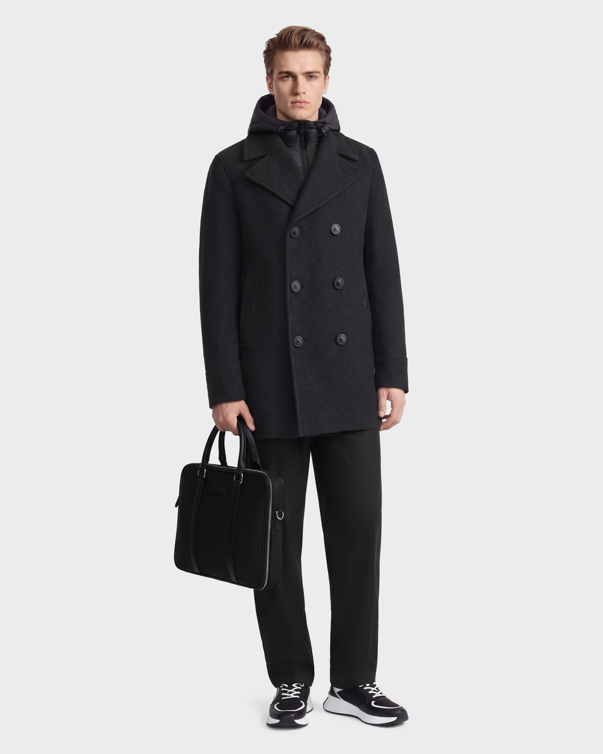 A man wears a charcoal LUCIEN - 6126508 Rudsak double-breasted wool-blend pea coat layered over a hooded jacket, black trousers, and sneakers while carrying a black briefcase.