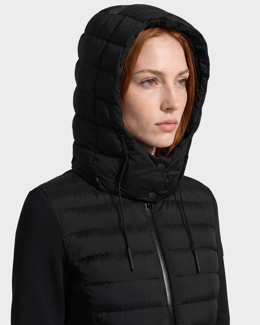 A woman with straight red hair wears the Rudsak MERINA - 8125056 Black quilted down jacket, which features a hood with drawstrings and is fully zipped up. She faces slightly to the right against a plain light background.