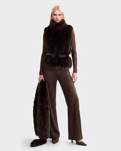 A person wears a brown ribbed mock-neck pullover and matching wide-leg trousers from the Rudsak MAULIN - 8524611 Mix Espresso set, styled with a dark brown faux fur vest cinched by a black belt while holding an additional matching fur garment.