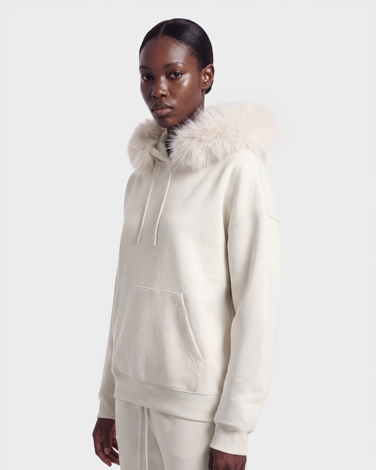 A person wears the Rudsak MILENA - 8526689 Vanilla hoodie, featuring a large, fluffy fur-trimmed hood, standing slightly turned against a plain light background.