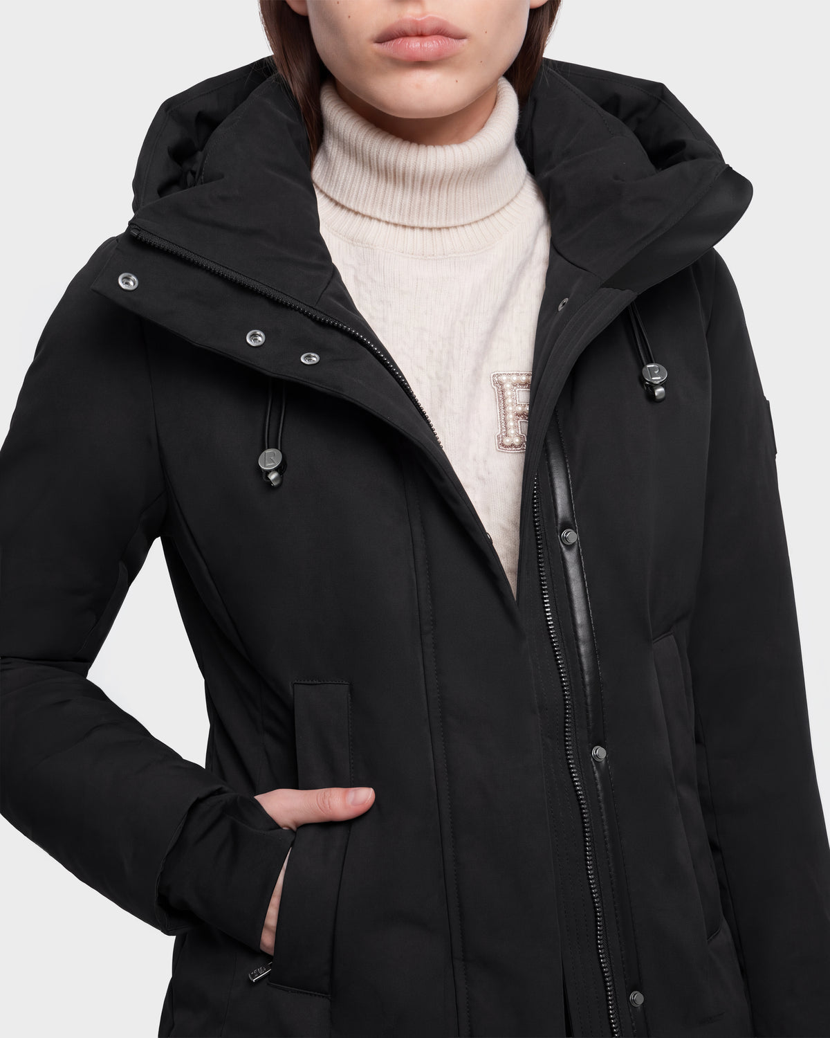 A person wearing the Rudsak OPAL - 8126618 Black hooded winter coat over a cream turtleneck sweater with an embroidered letter E, standing with one hand in the coat pocket.