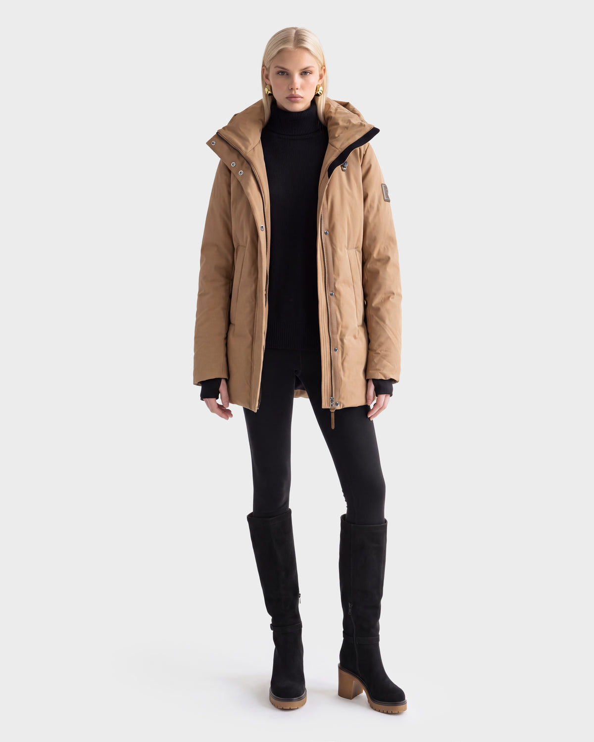 A woman models the Rudsak OPAL - 8126618 Camel parka, paired with a black turtleneck, leggings, and heeled boots, standing facing forward against a plain white background.