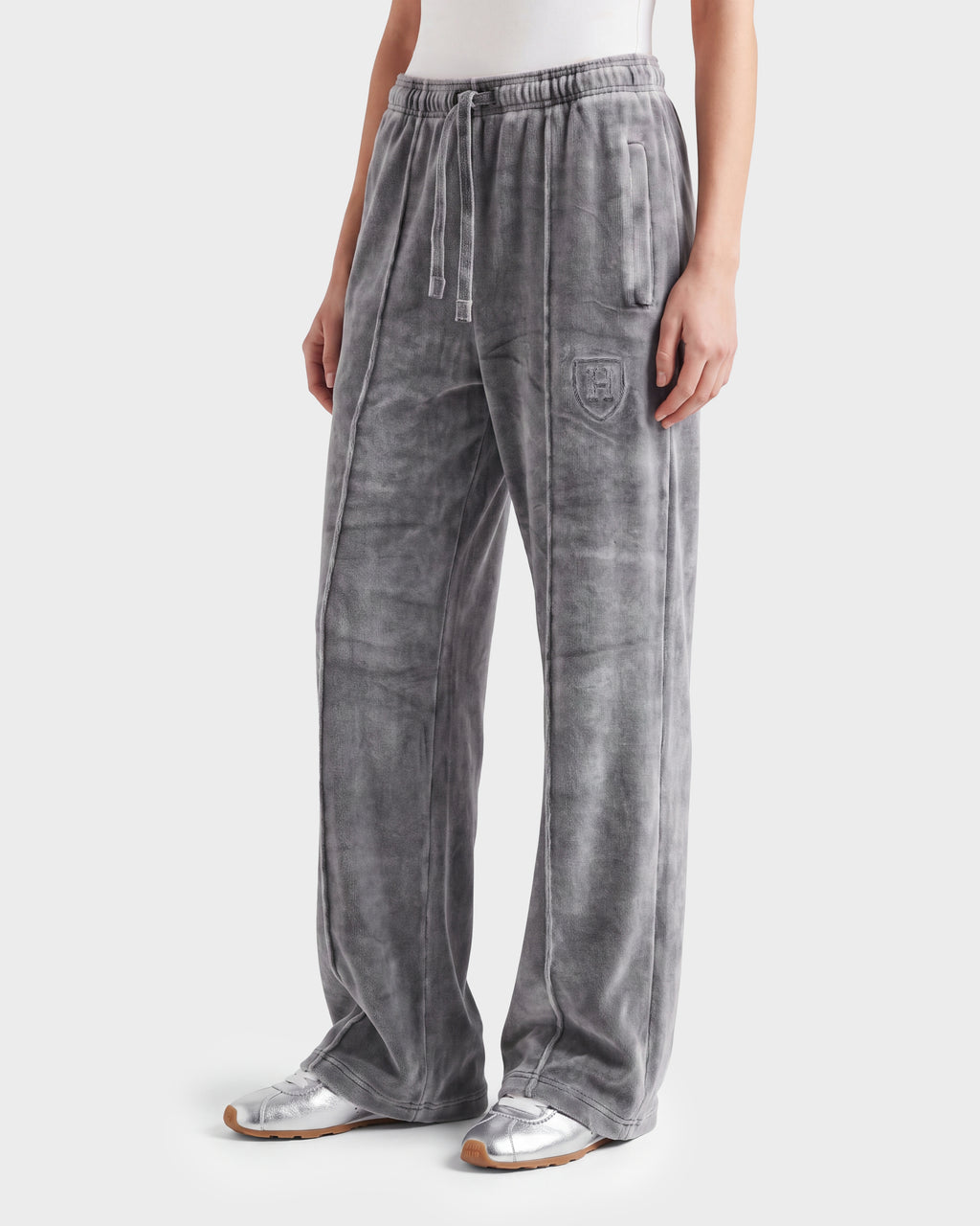 A person wears the Rudsak OLIVYA HV - H526903 Titanium gray velour sweatpants featuring a drawstring waist, side pockets, and an embroidered crest on the left thigh, styled with a white top and silver sneakers.