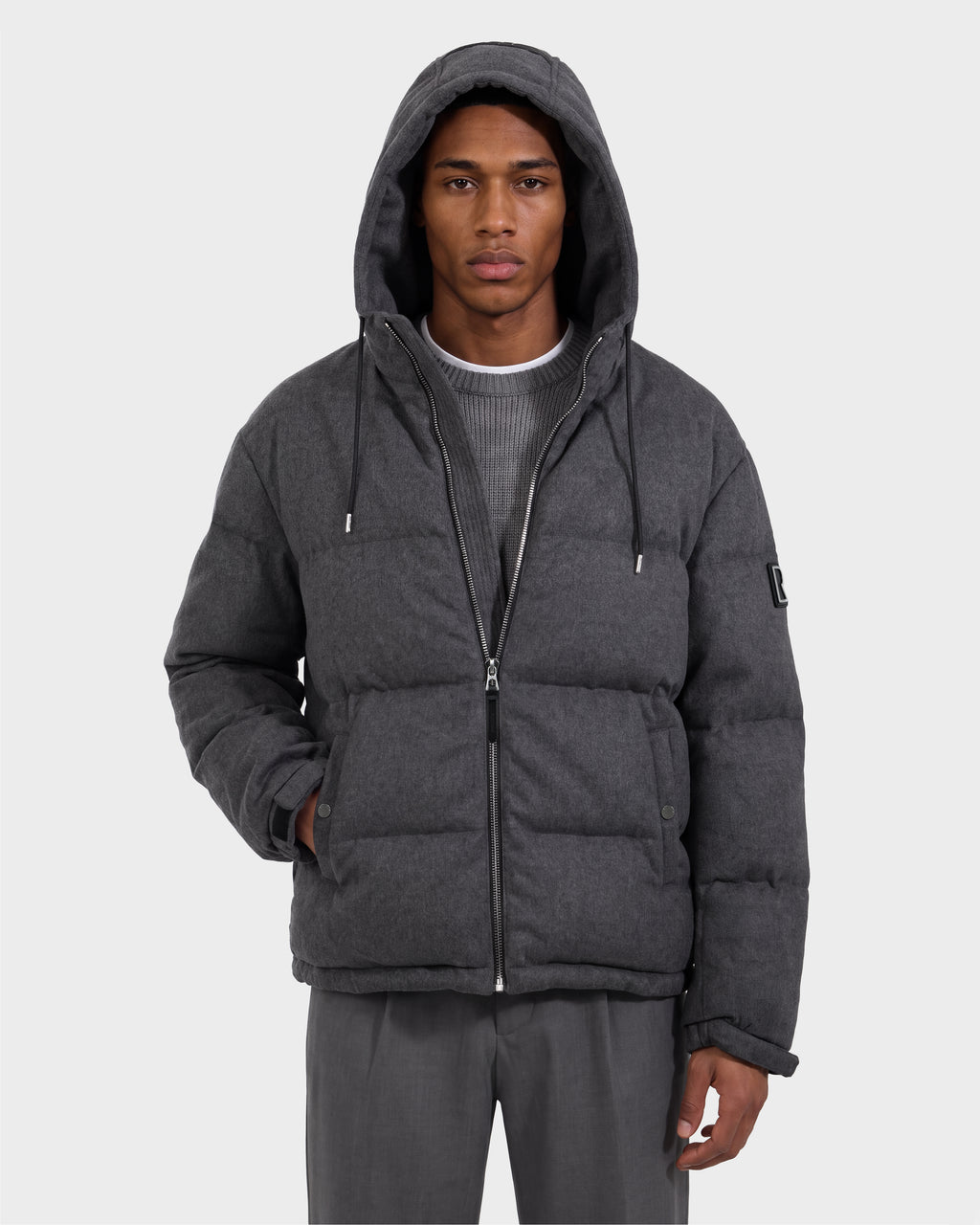 A person wearing the Rudsak STERLING W - 6126760 Heather Charcoal hooded puffer jacket stands with one hand in their pocket, layered over a gray sweater and gray pants, facing forward against a plain light background.