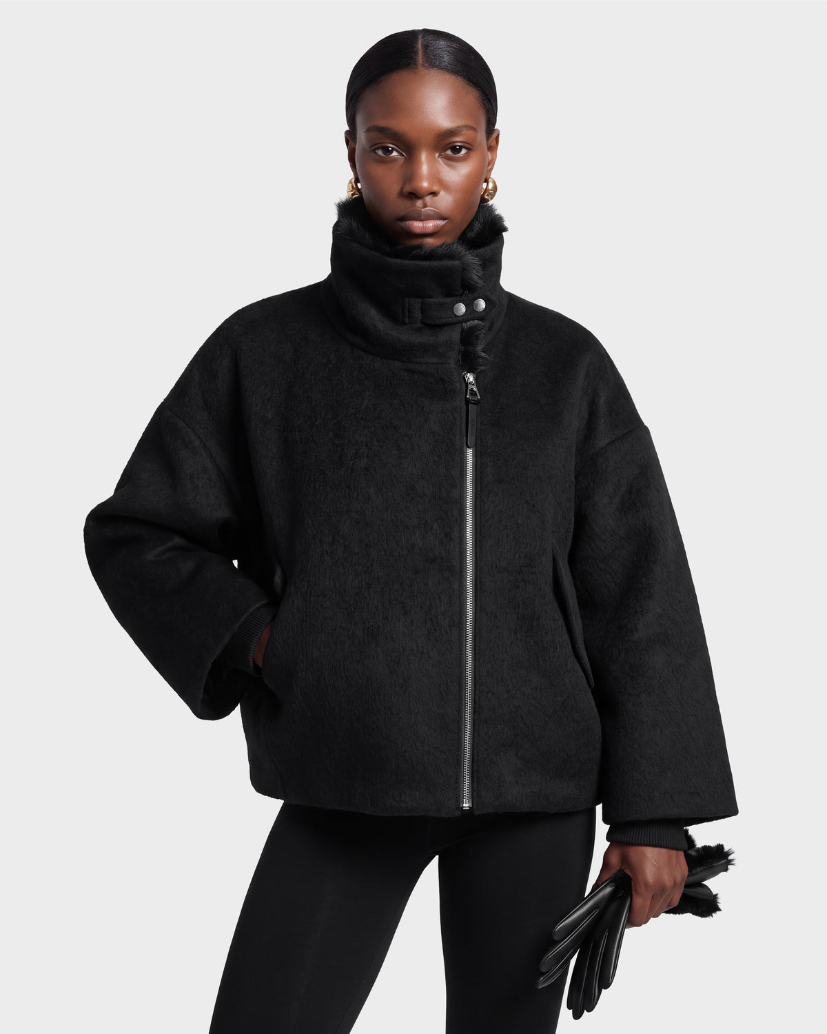 A woman wears the Rudsak ZARIA - 8126999 Black high-neck jacket and matching black leggings, holding black gloves while standing against a plain light gray background.