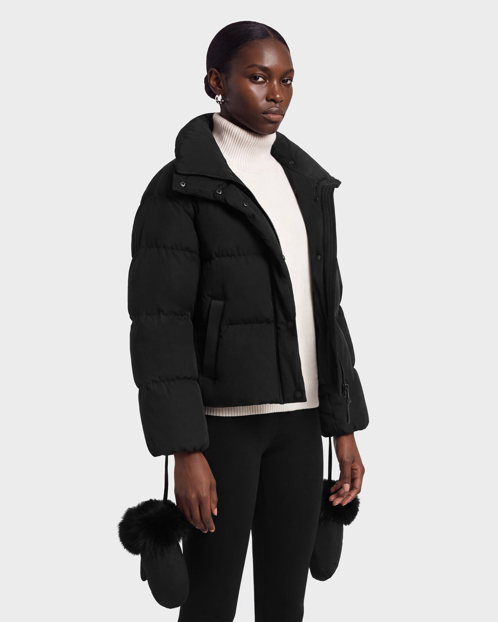 A person wears a black ALONDRA - 8126992 Black cropped puffer jacket by Rudsak over a white turtleneck, with matching black mittens featuring fur trim hanging from the sleeves.