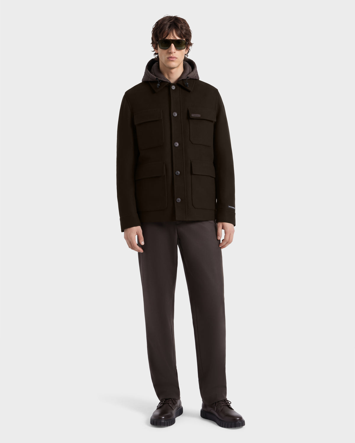 A man wearing the Rudsak BRUNO - 6126716 Espresso dark brown hooded button-up jacket, dark trousers, and dark sunglasses.