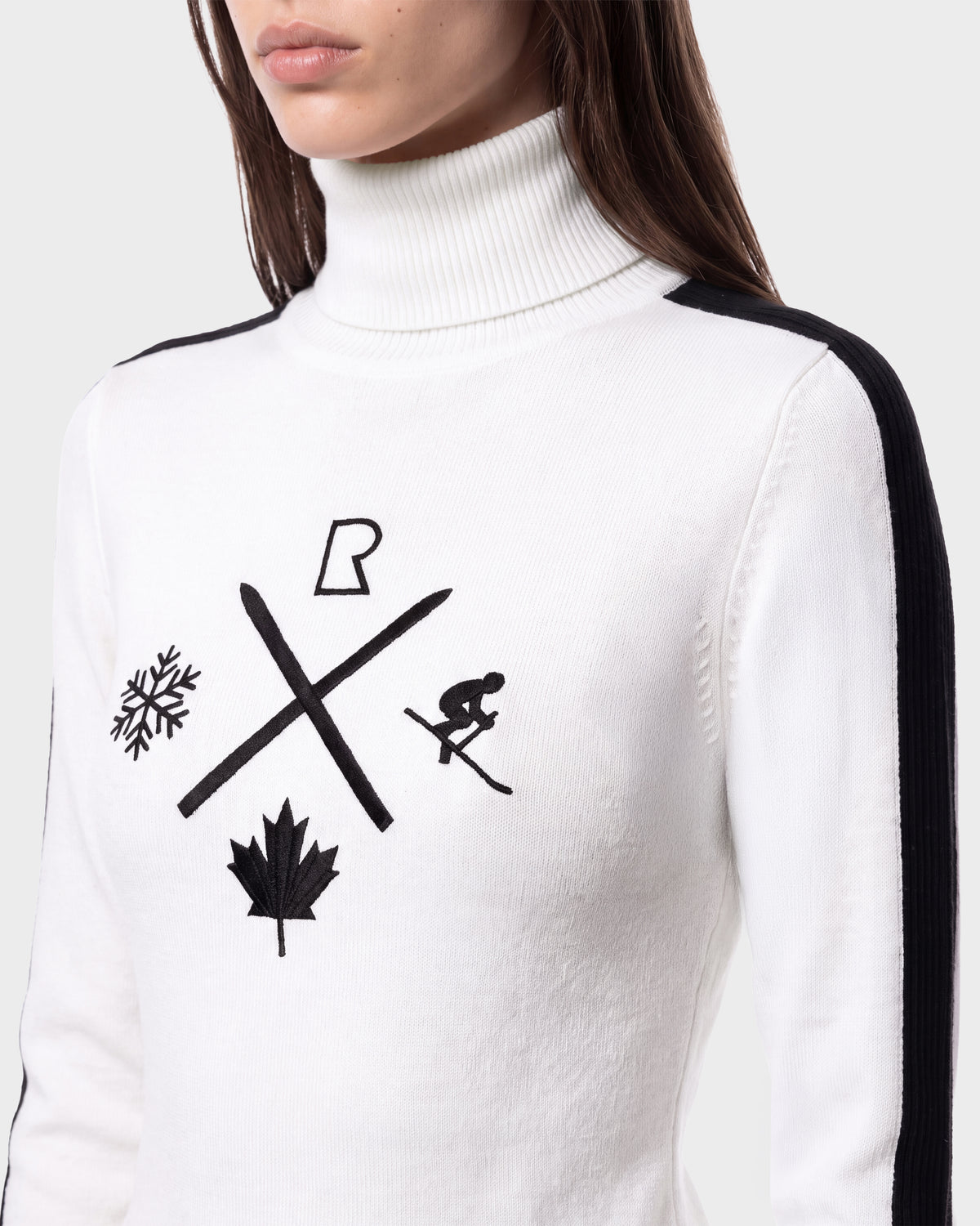 A woman wearing the ELIZABETH - 8524623 Optic White women’s ski turtleneck sweater from Rudsak, featuring black shoulder stripes and bold black ski-themed graphics—including skis, a skier, a snowflake, and a maple leaf—on the front; perfect for elevating your winter wardrobe or relaxing après-ski.