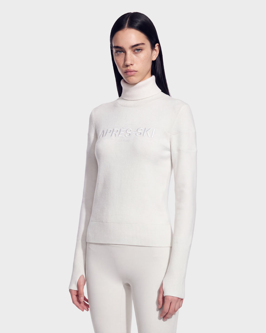 A woman with long dark hair wears the ERIKA - 8525705 Iceland white turtleneck sweater by Rudsak, an essential for any winter wardrobe featuring APRÈS-SKI text, paired with matching white pants and standing against a plain light background.