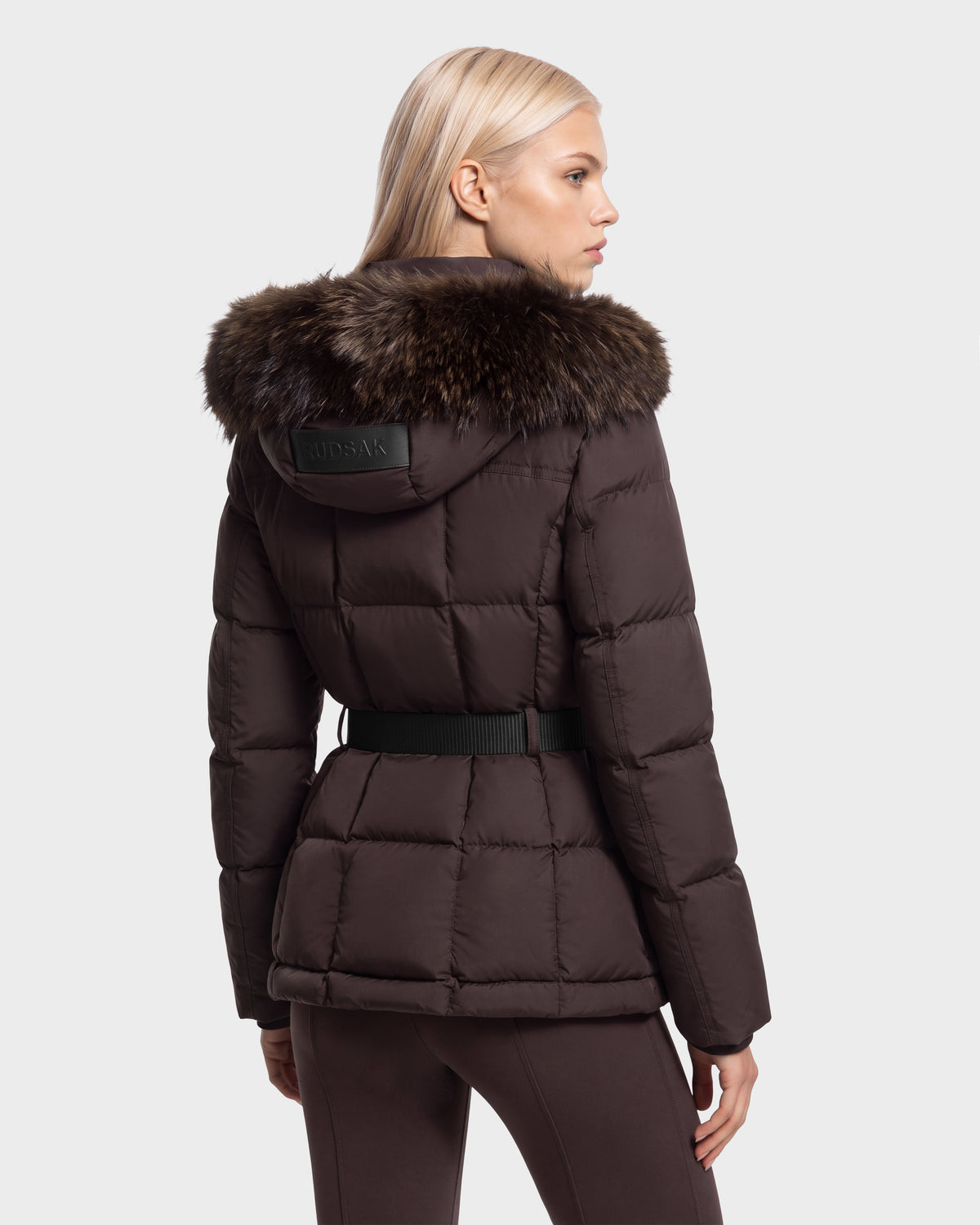 A woman seen from behind wearing a Rudsak GRACE FF - 8126535 Espresso dark brown, quilted puffer jacket with a fur-trimmed hood and a black waist belt.