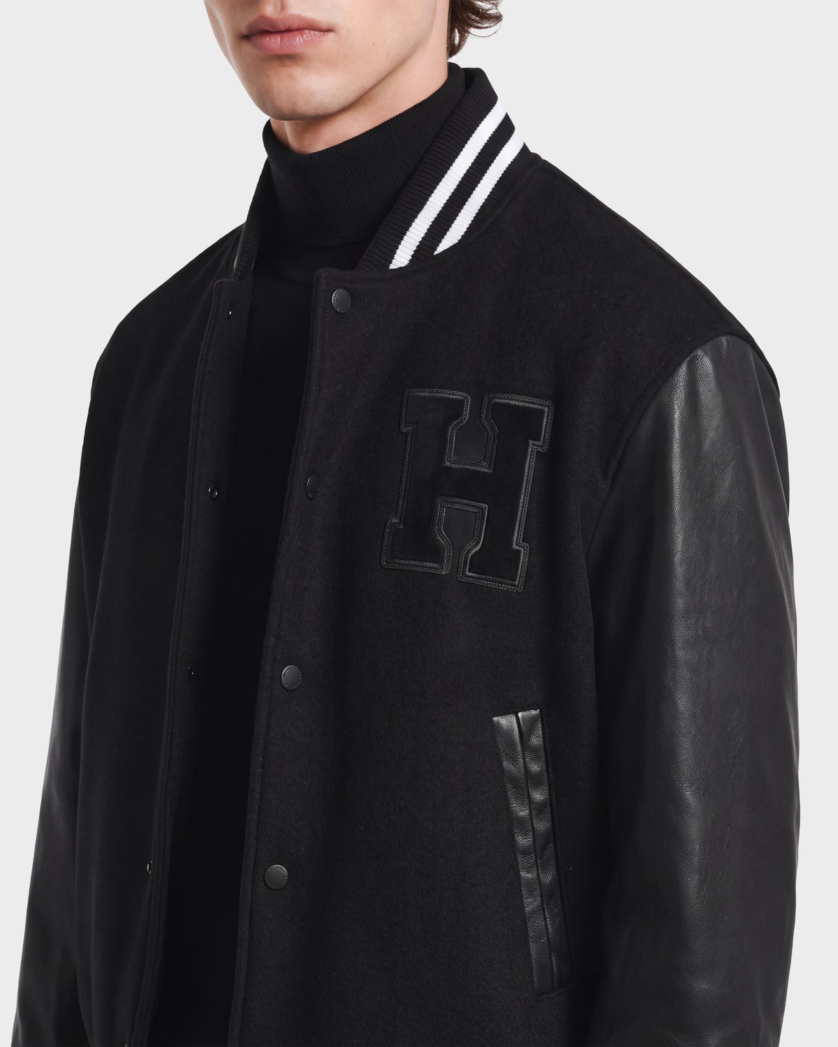 A person wearing the JOHN HV - H125504 Black collegiate jacket by Rudsak—a black varsity jacket with leather sleeves, a striped collar, and a large H patch on the chest—layered over a black turtleneck. Only the lower face and torso are visible.