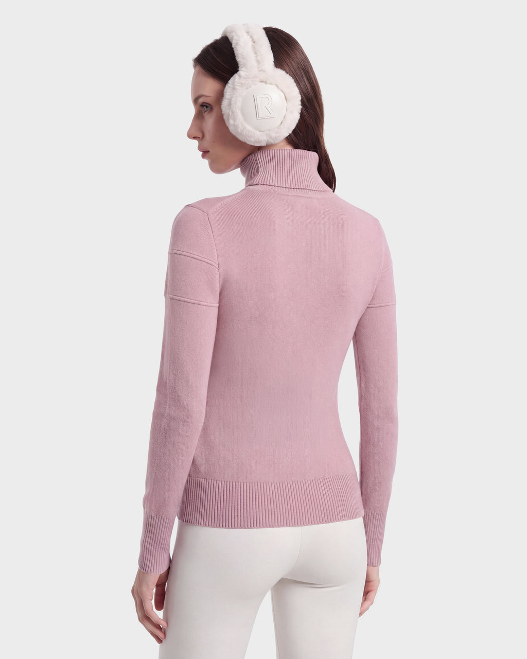 A woman facing away is wearing the ERIKA - 8525705 Vintage Rose turtleneck sweater by Rudsak, paired with white pants and white quilted earmuffs featuring the R logo, set against a plain light background.