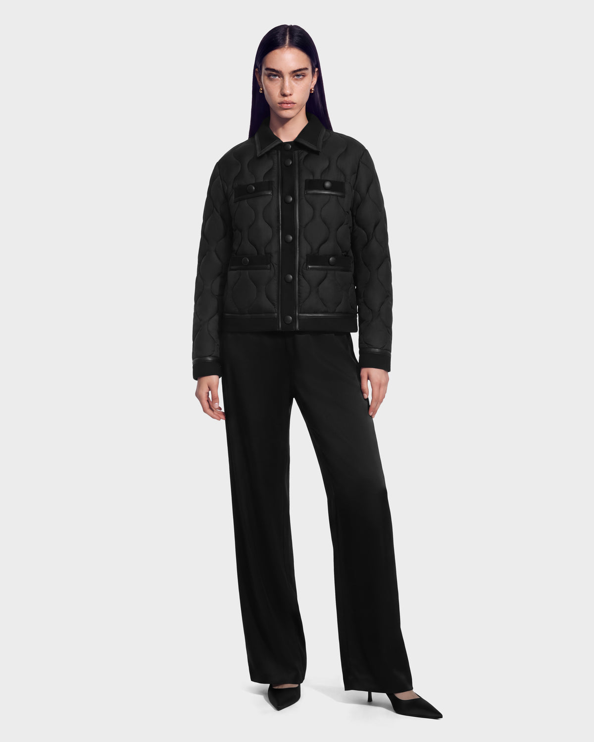 A person wearing a black JOLIE - 8125036 Black quilted down jacket by Rudsak with leather-look trim over black trousers and pointed-toe heels.