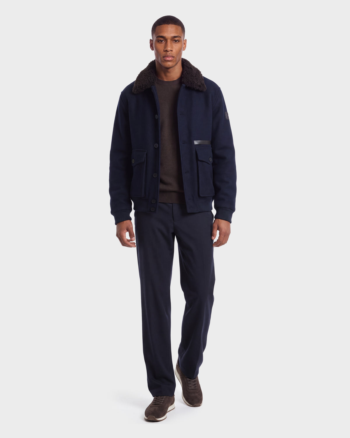 A man wearing a Rudsak JULIUS - 6126723 Navy wool-blend bomber jacket with a dark faux-fur collar over a brown sweater and matching navy trousers.