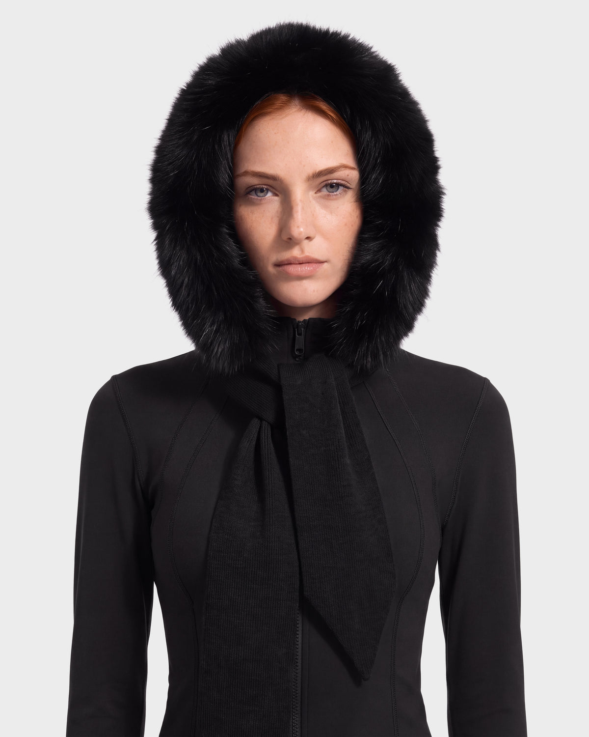 A woman wears the Rudsak KARYMA - 8426507 Black jacket, featuring a large plush trim hood-and-scarf hybrid, as she looks directly at the camera with a neutral expression against a plain background—a standout winter accessory.