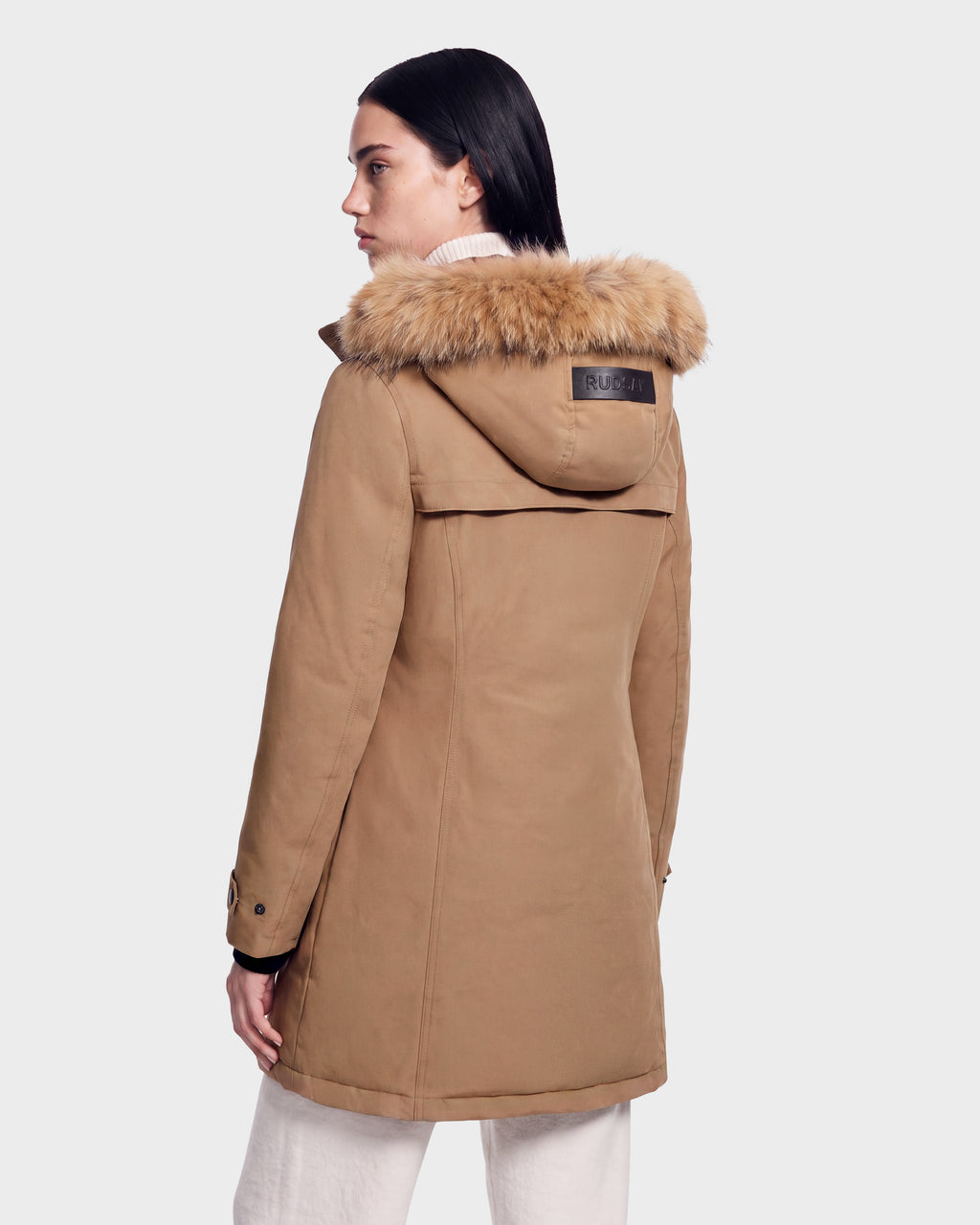 A woman wears the Rudsak KEYSHIA - 8126517 Camel down coat with a fur-trimmed hood and visible back label, facing away and looking left against a plain light background.