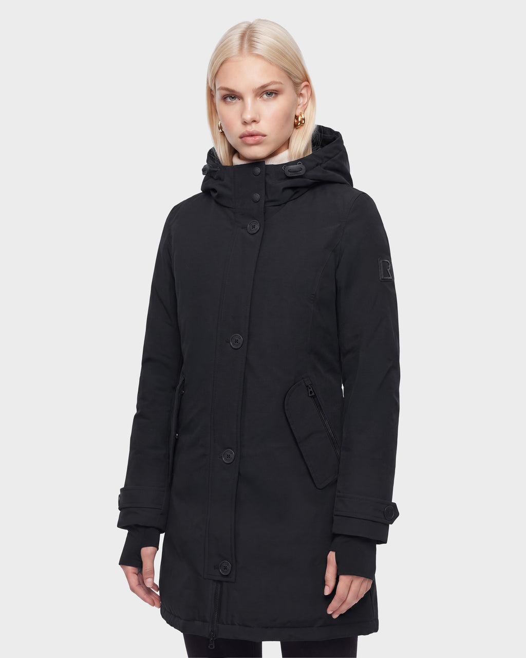 A woman with straight blonde hair models the Rudsak KEYSHIA - 8126517 Black winter coat, featuring large buttons and zippered pockets, standing against a plain white background.