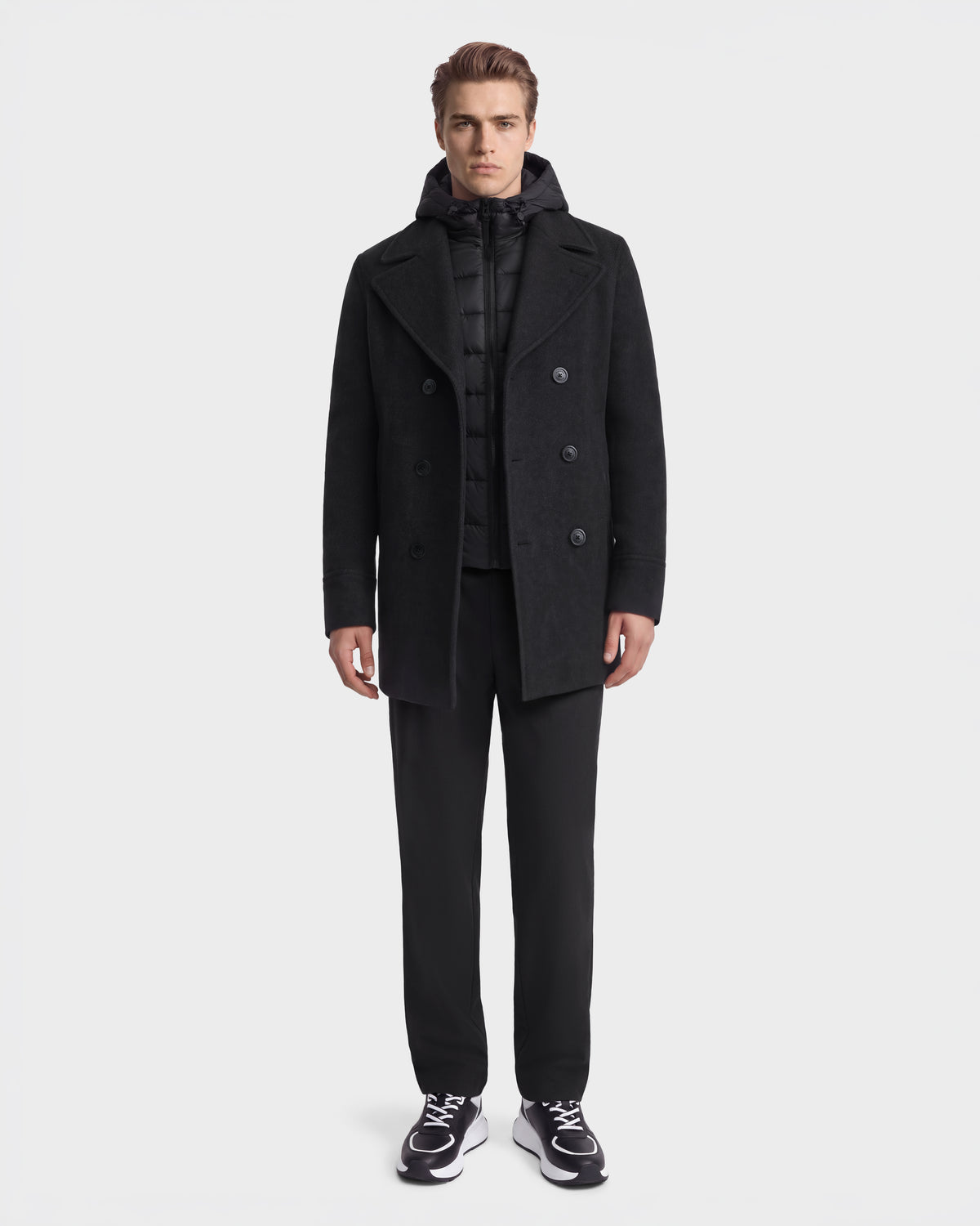 A man wearing a Rudsak LUCIEN - 6126508 Charcoal wool-blend pea coat over a black duck down quilted hooded vest, black trousers, and black and white sneakers.