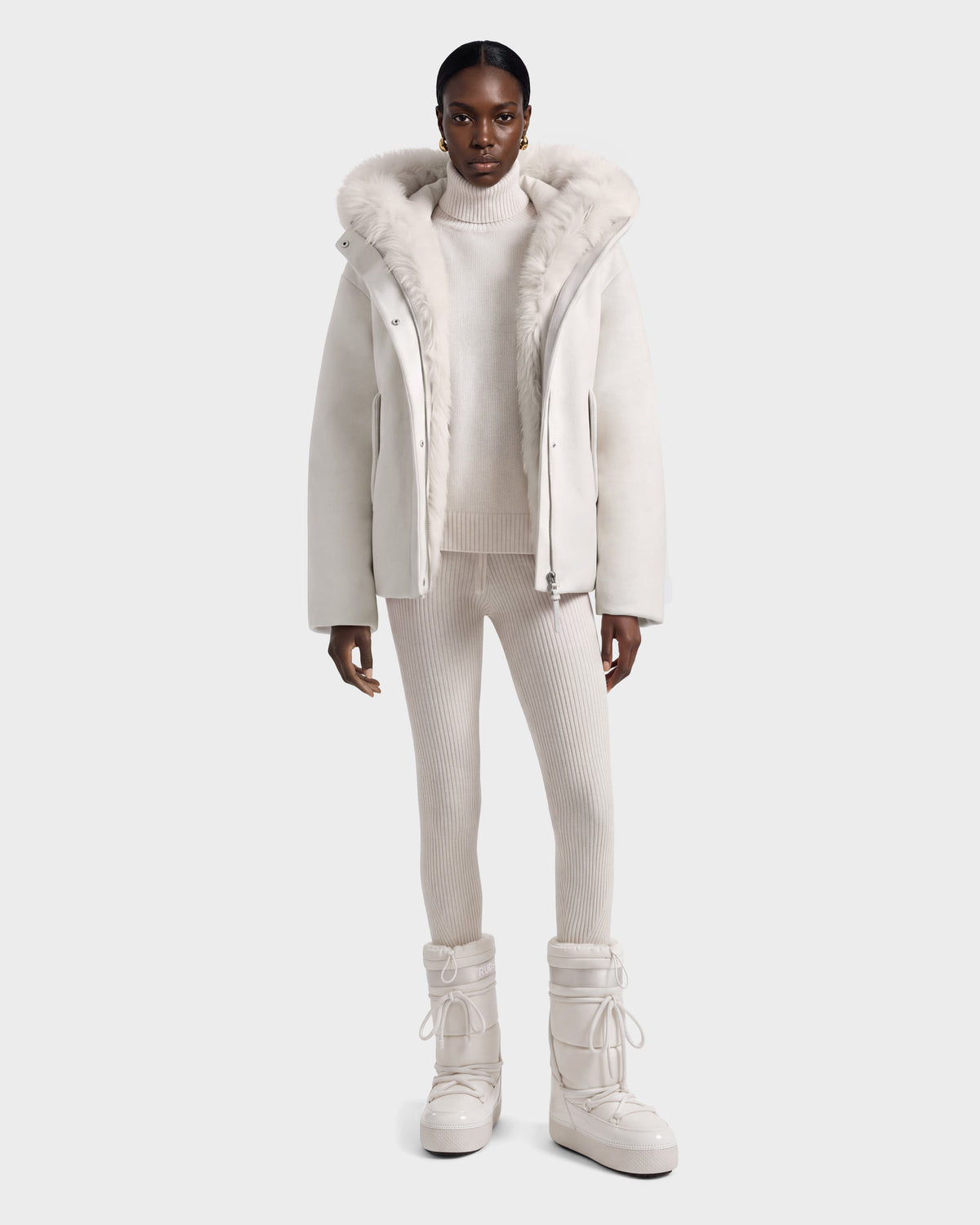 A person wearing a HAZEL - 8126981 Vanilla white hooded winter coat with faux-fur trim, a matching turtleneck sweater, ribbed leggings, and lace-up snow boots.