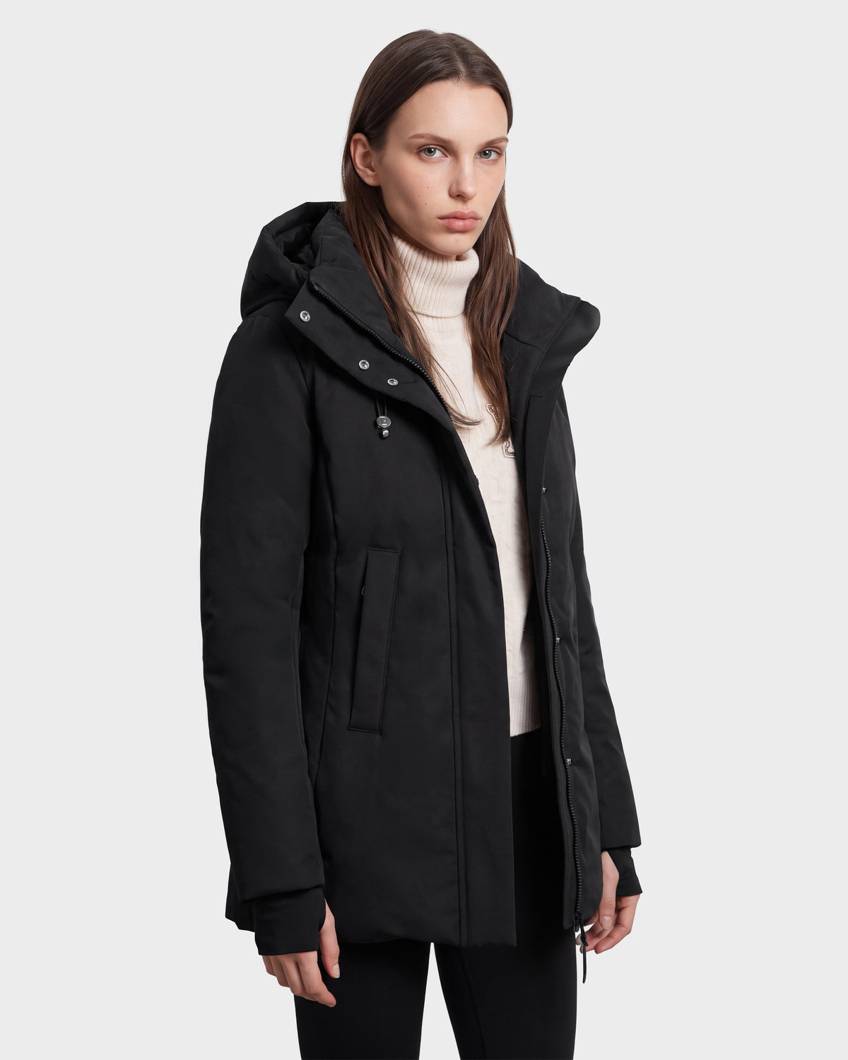 A woman wearing the Rudsak OPAL - 8126618 Black hooded winter coat over a white turtleneck sweater stands against a plain light background, looking at the camera with a neutral expression.