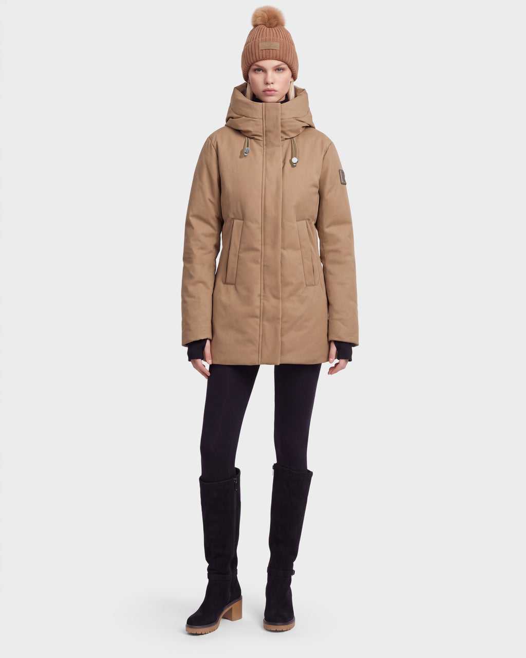 A woman stands facing forward against a white background, wearing the Rudsak OPAL - 8126618 Camel parka, black leggings, knee-high black boots, and a tan knit hat with a pom-pom.