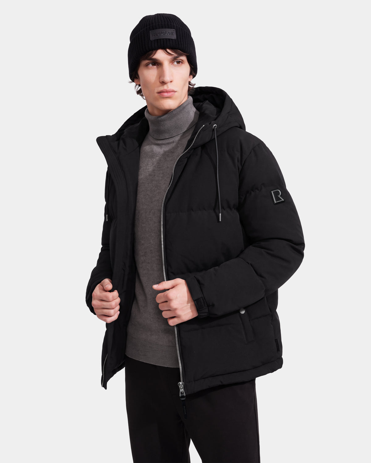 A person wearing the Rudsak STERLING EL - 6126759 Black puffer jacket, paired with a gray turtleneck and black knit beanie, stands against a plain light background, gazing slightly to the side with one hand touching the jacket.