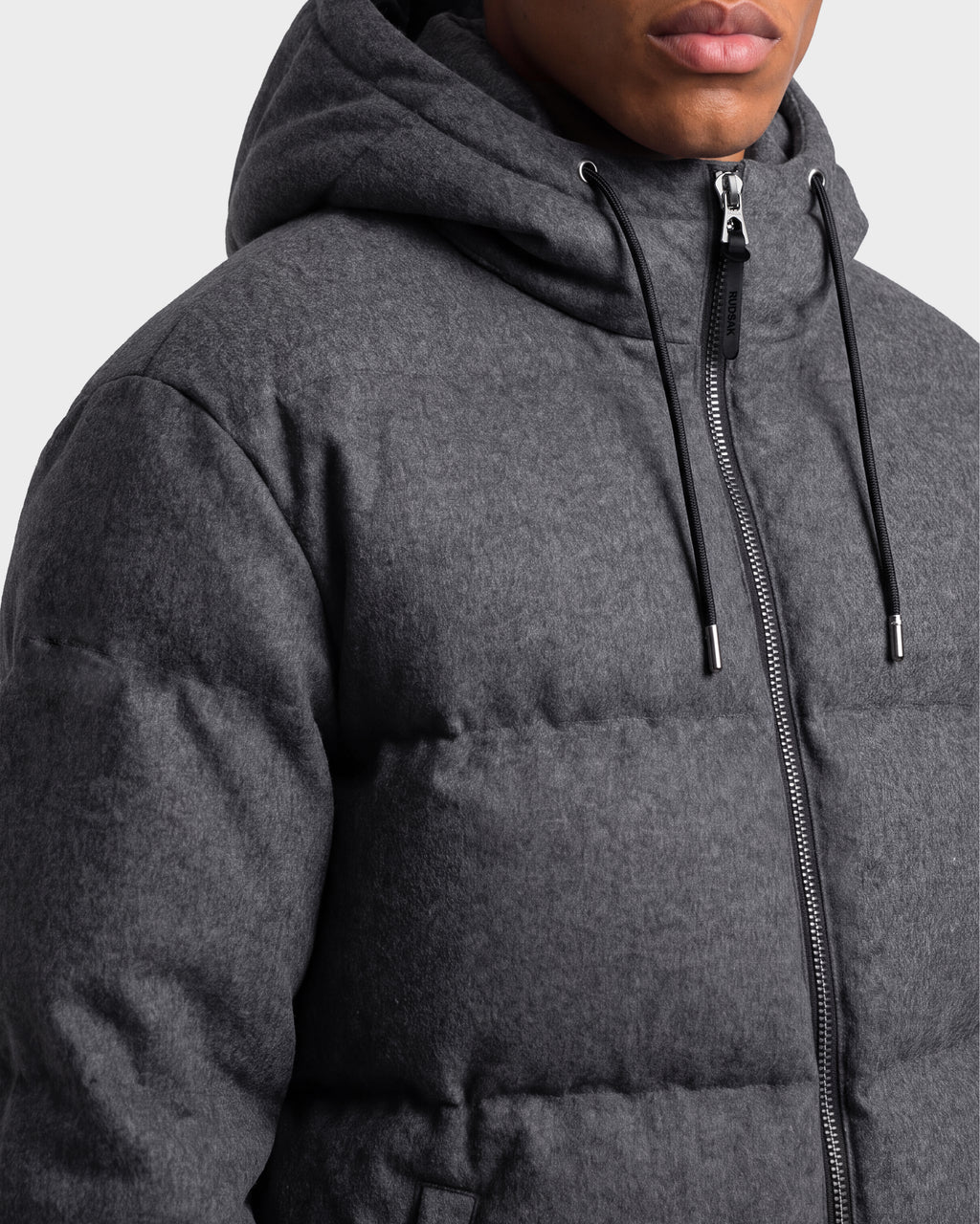 A person is shown from the shoulders to the lips against a plain background, wearing the Rudsak STERLING W - 6126760 Heather Charcoal hooded puffer jacket featuring a zipper and drawstrings.
