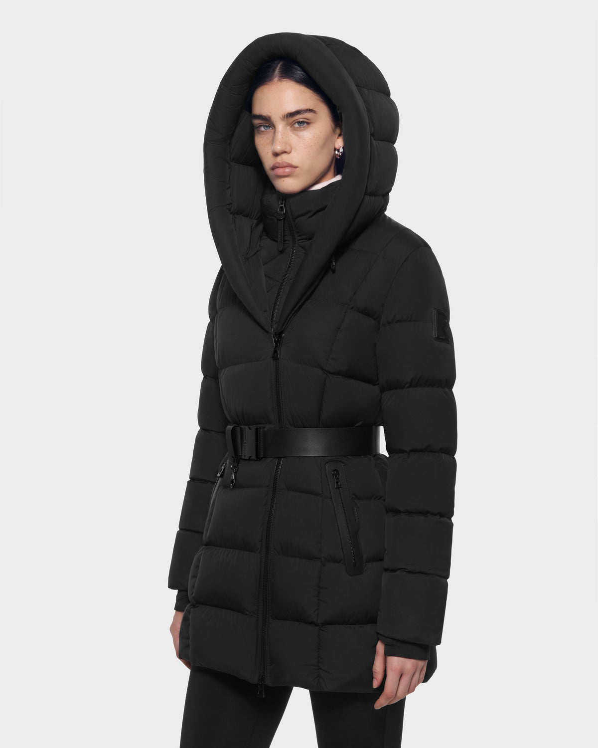 A person wearing a black Rudsak TAYLOR M - 8125965 quilted winter coat featuring a hooded design, a belted waist, and zippered pockets.