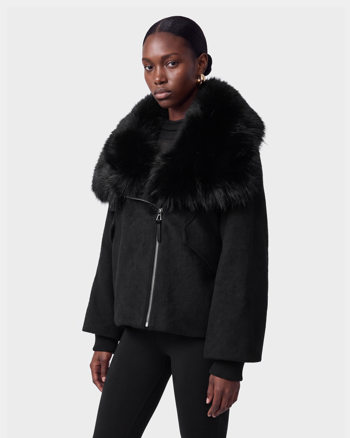 A woman wearing the Rudsak ZARIA - 8126999 Black faux fur-collared jacket with a zipper, paired with black leggings and gold hoop earrings, stands against a plain light gray background.