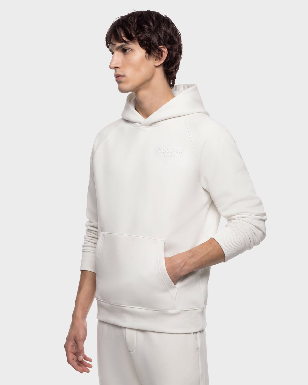 A man wearing a RUDSAK BRIAR - 9525001 Vanilla M cream-colored hooded sweatshirt with a front kangaroo pocket.