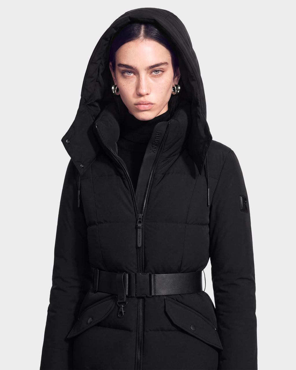 A person with straight dark hair, wearing the Rudsak ADELINE - 8124544 Black quilted winter jacket with a belt and silver hoop earrings, stands facing forward against a plain light background.