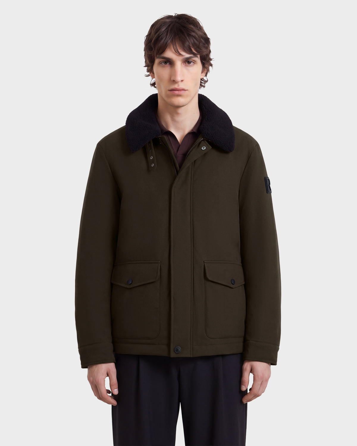 A man wearing a Rudsak ANTOINE - 6126506 Olive insulated winter jacket with an olive green finish, a dark faux-fur collar, and two large front flap pockets.