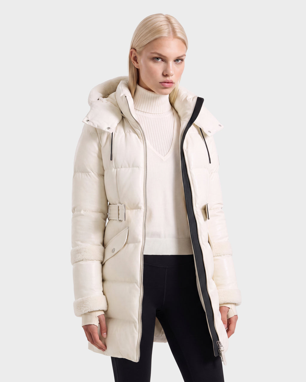 A person wearing a Rudsak AUDREY L - 8124585 Vanilla ivory hooded down puffer jacket with a belted waist and shearling-lined trim on the cuffs and hood.