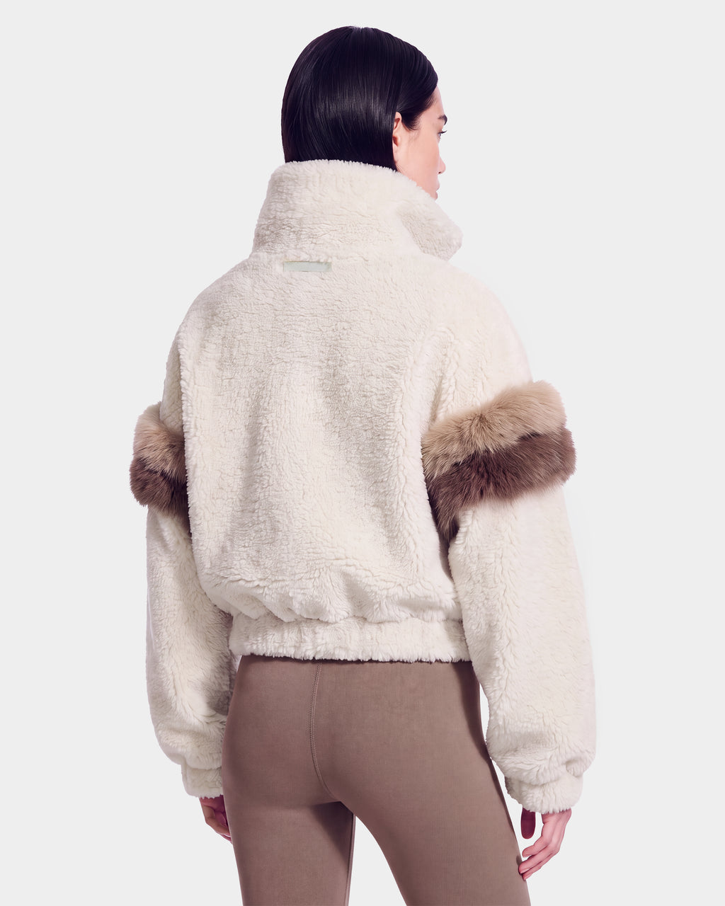 A person is shown from the back wearing the Rudsak ALEIA - 8526693 Vanilla, a white, fluffy faux-fur sweater with a high collar and brown fur accents on the sleeves. They have short dark hair and are also wearing fitted, light brown pants.