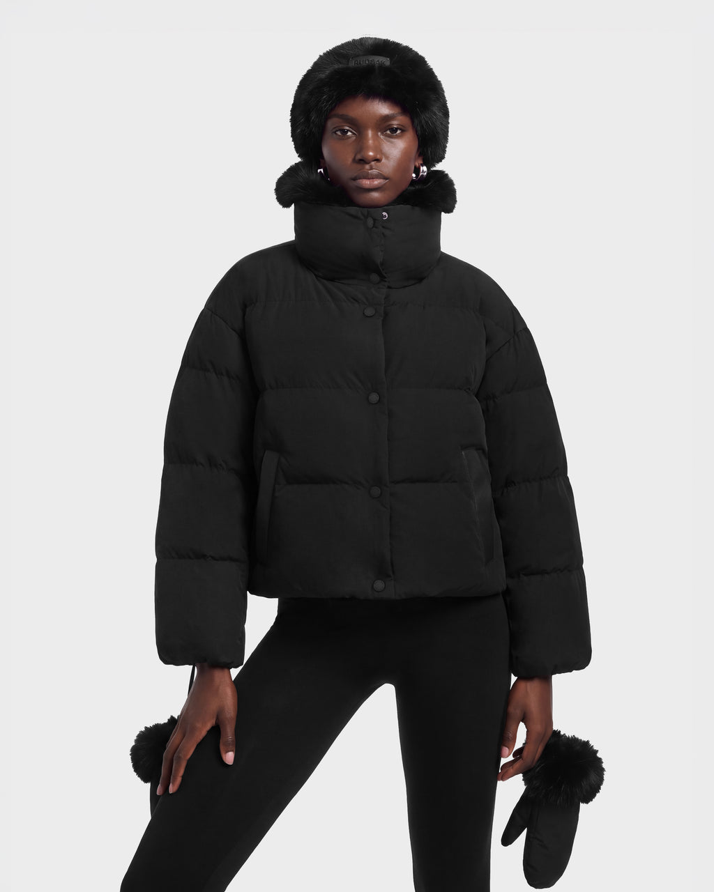 A person wears a black ALONDRA - 8126992 Black, cropped puffer jacket with a high collar, matching faux fur-trimmed hat, and mittens made with RDS-certified down.