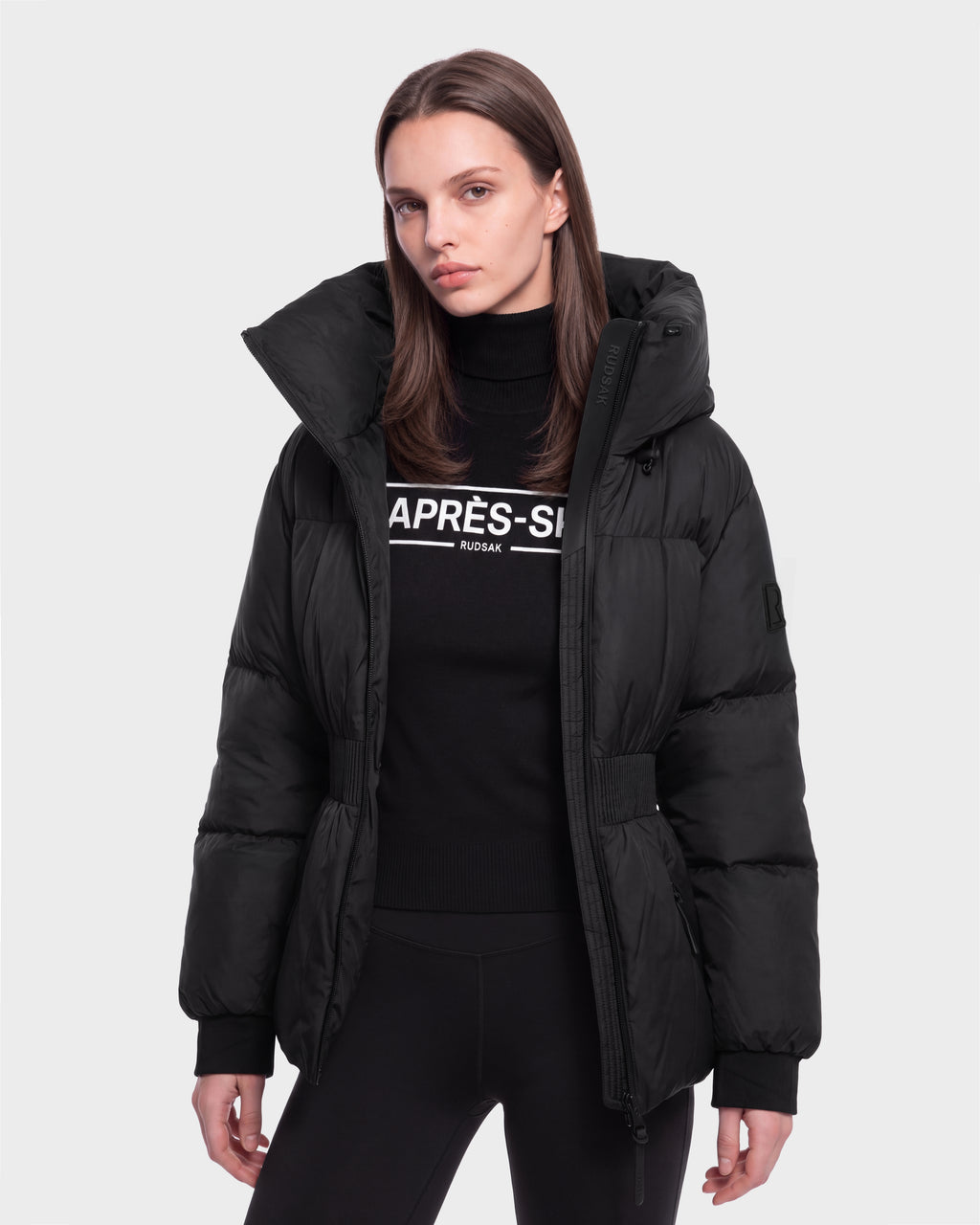 A woman with straight brown hair wears the Rudsak DIAMOND - 8126772 Black puffer jacket over a black APRÈS-SKI turtleneck, standing against a plain light gray background.