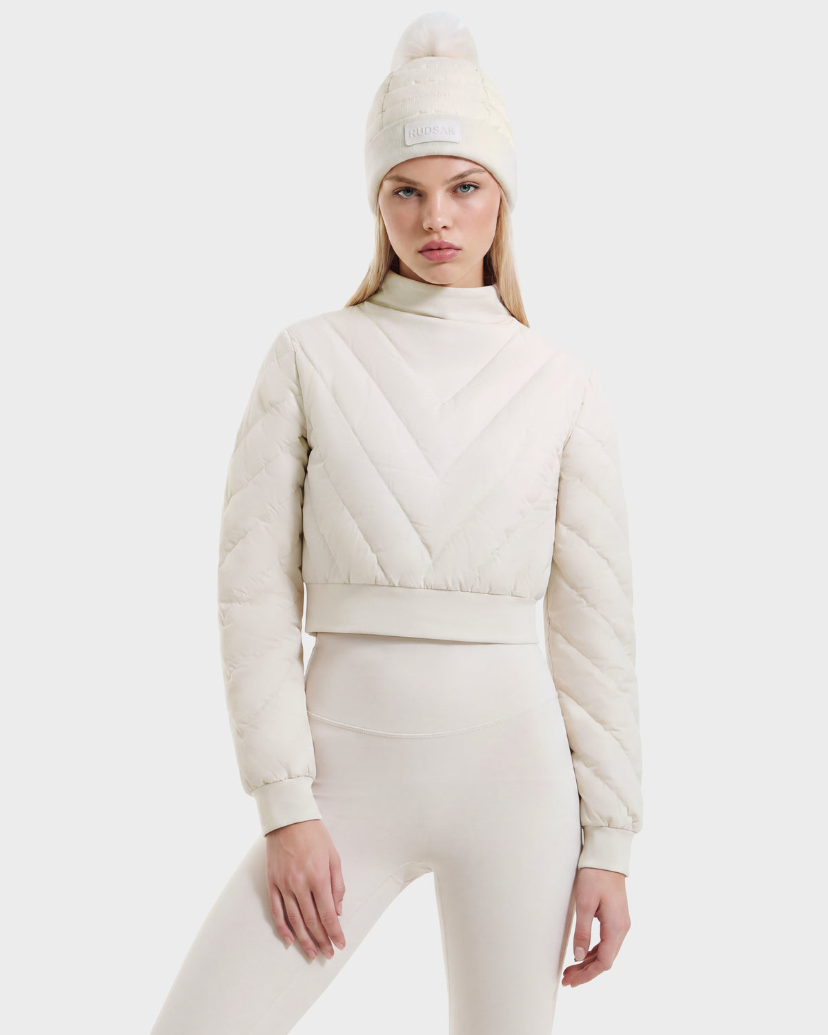 A person wears a cream-colored, RDS-certified insulated KATYA - 8526531 Vanilla crop puffer jacket and a matching pom-pom beanie as a stylish après-ski layer.