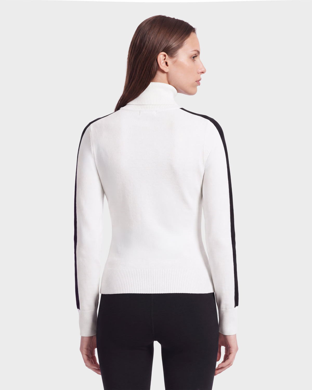 A woman with straight brown hair wears the Rudsak ELIZABETH - 8524623 Optic White women’s ski turtleneck sweater, featuring black stripes on the sleeves, paired with black pants as she faces away from the camera against a plain background—an effortlessly chic addition to any winter wardrobe.