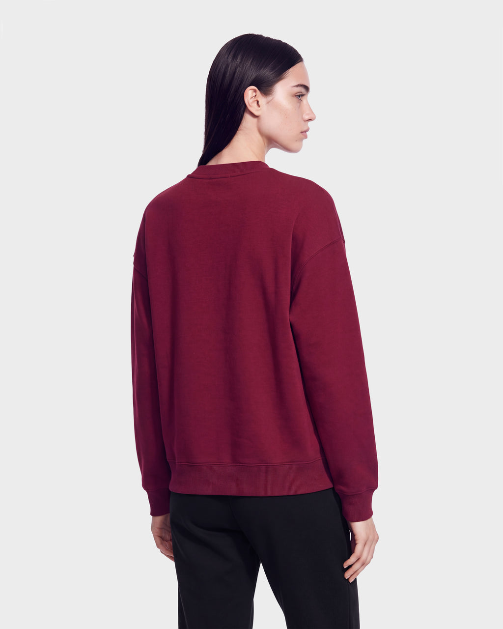 A person with long dark hair is wearing the Rudsak HARVARD ONE CRIMSON SWEATSHIRT 9524507 and black pants, standing and facing away to the left against a plain light background.