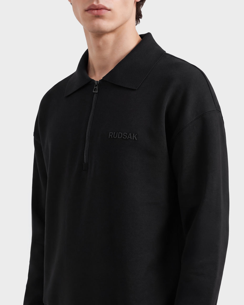 A person wearing the Rudsak IDRIS - 6526500 Black zip-collar sweatshirt with a relaxed fit and logo embroidery, shown from shoulders to lips against a plain background.