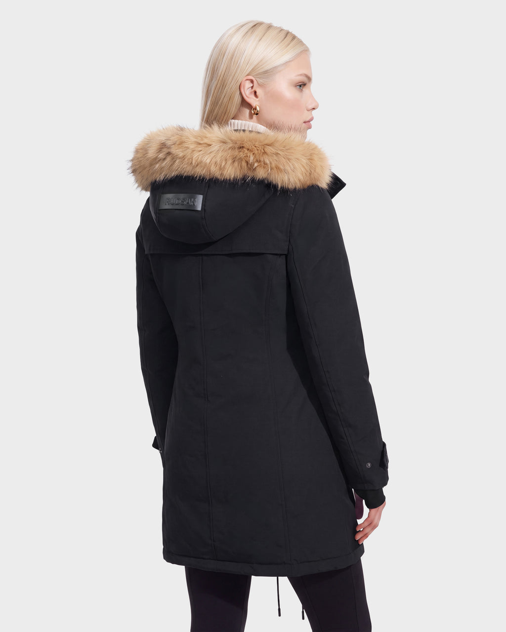 A woman with blonde hair wears the Rudsak KEYSHIA - 8126517 Black coat with a fur-trim hood, seen from the back against a plain light background—a cold-weather essential.