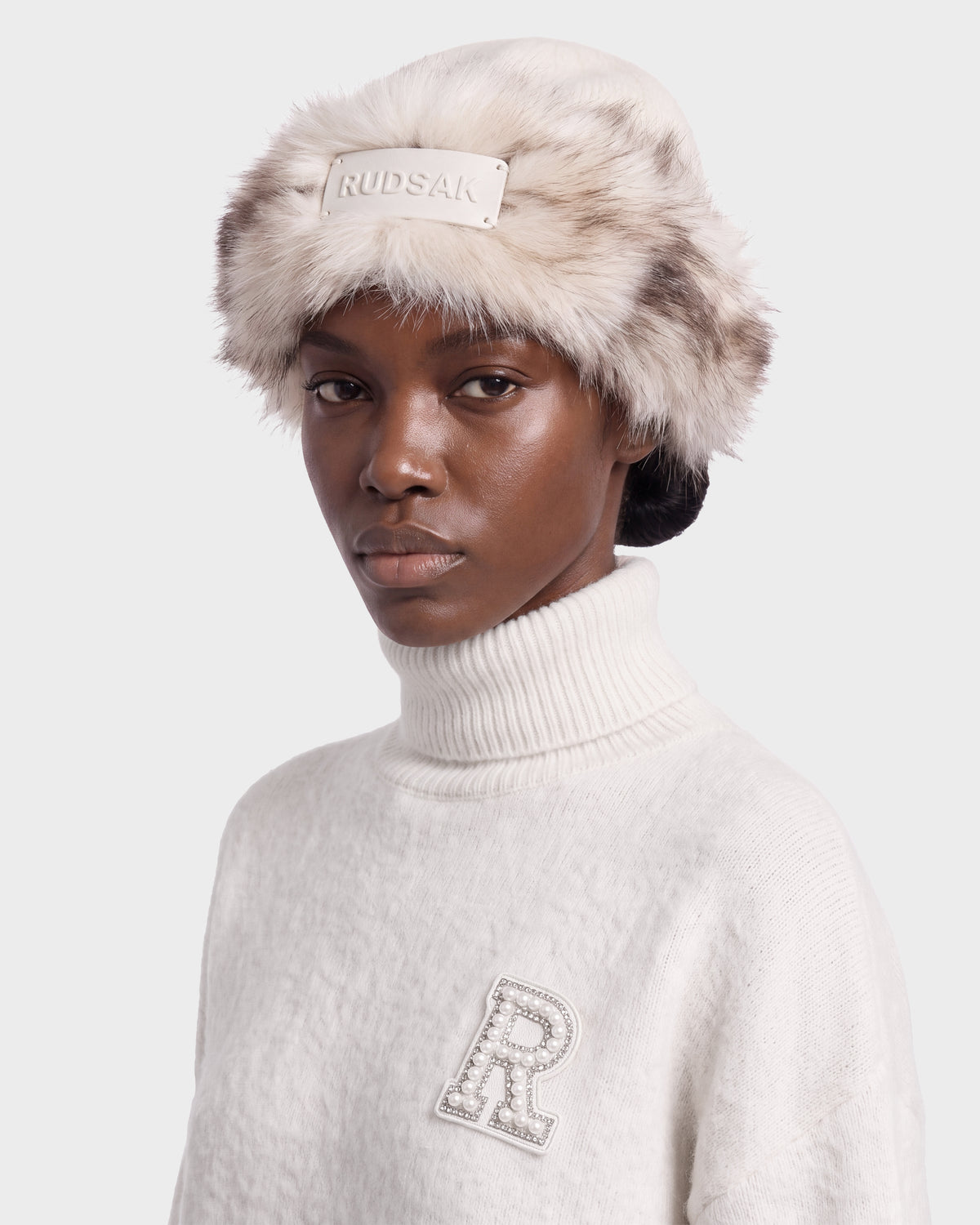 A person wearing a LORETTA W - 8426510 Vanilla white turtleneck sweater with a rhinestone-embellished R patch and a RUDSAK faux fur hat featuring a rectangular logo tag on the front.