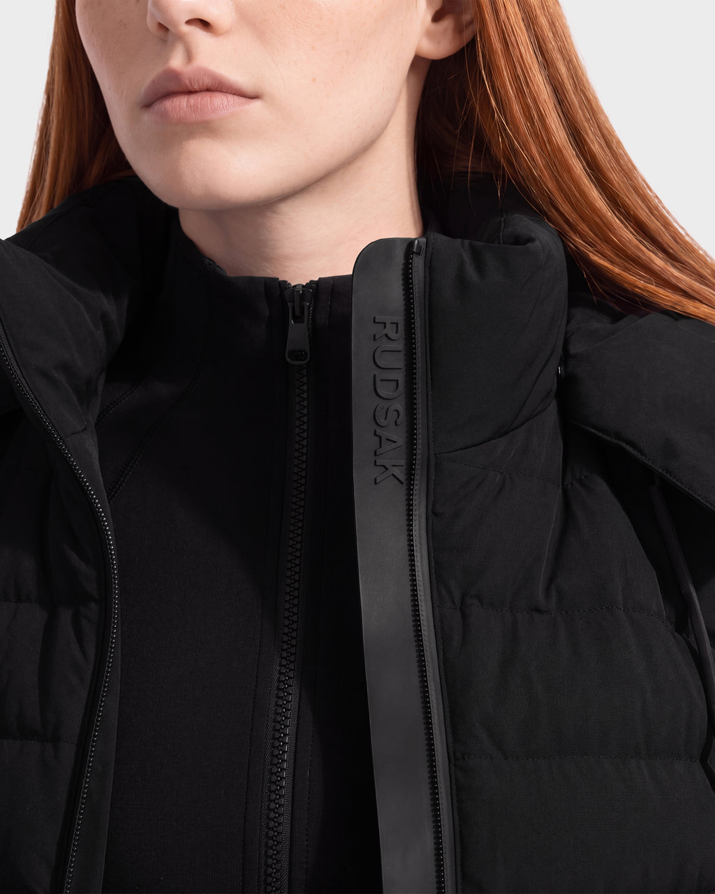 A woman with long red hair is shown from the shoulders up, facing slightly left, wearing the Rudsak MERINA - 8125056 Black women’s light jacket. The jacket features a high collar and visible RUDSAK branding on a leather strip near the zipper.