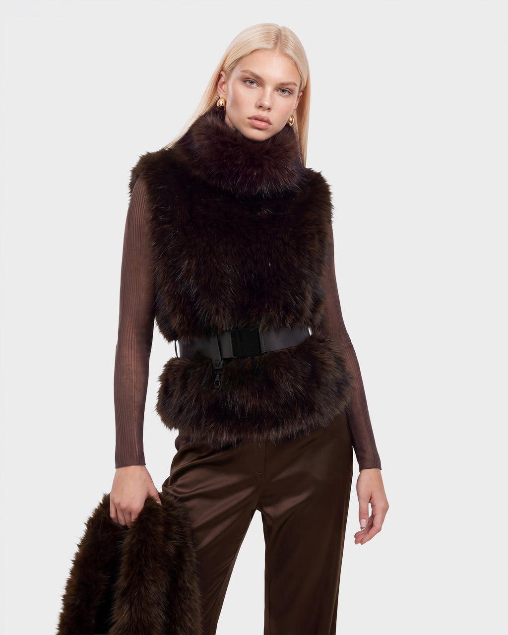 A model wearing a MAULIN - 8524611 Mix Espresso dark brown faux fur vest with a black belted waist over a long-sleeved mock-neck pullover and trousers, holding a matching fur accessory.
