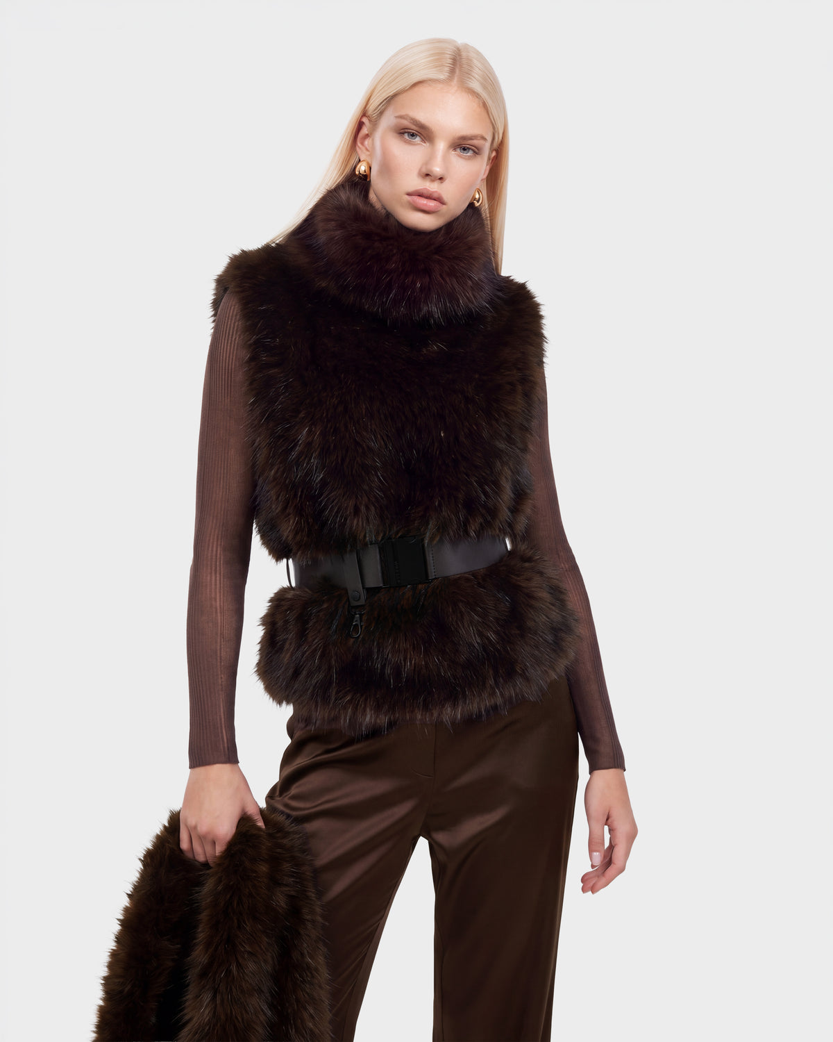 A model wearing a MAULIN - 8524611 Mix Espresso dark brown faux fur vest with a black belted waist over a long-sleeved mock-neck pullover and trousers, holding a matching fur accessory.